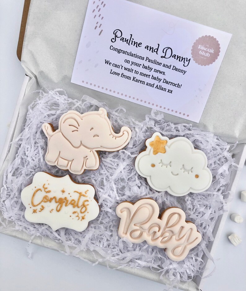 May include: A gift box filled with white shredded paper and four decorated cookies. The cookies are decorated with gold and white icing and feature a baby elephant, a cloud with a star, the word "Congrats" and the word "Baby". The box also includes a card with the text "Pauline and Danny Congratulations Pauline and Danny on your baby news. We can't wait to meet baby Darroch! Love from Karen and Allan xx" and a small sticker that says "Biscuit Club".
