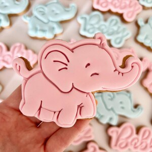 May include: A pink elephant cookie with a smiling face. The cookie is decorated with white icing and is being held in a hand.