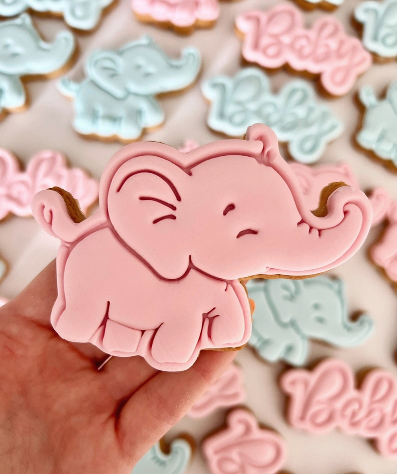 May include: A pink elephant cookie with a smiling face. The cookie is decorated with white icing and is being held in a hand.