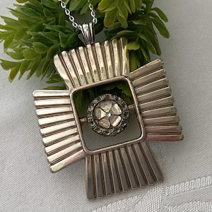 May include: Silver-toned pendant necklace with a cross-shaped design. The pendant features a square center with a circular, gem-studded element. The cross has a ridged, sunburst-like pattern. The necklace is displayed on a white surface with green foliage.