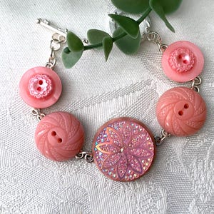 May include: A pink button bracelet with a silver chain. The bracelet features various sizes and styles of pink buttons, including a large button with a floral design. The buttons are connected by a silver chain and clasp.
