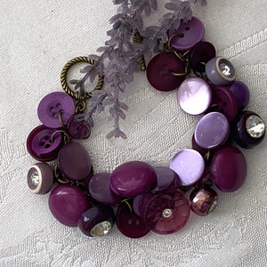May include: A handmade bracelet featuring a variety of purple buttons in different shades and sizes. Some buttons have a shiny, pearlescent finish, while others are adorned with rhinestones. The bracelet is accented with a sprig of lavender.