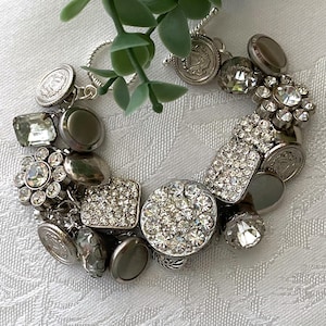 May include: A silver-toned charm bracelet featuring a variety of circular and rectangular components, including clear faceted crystals, smooth metallic discs, and textured coin-like charms, all connected by a silver chain with a toggle clasp.