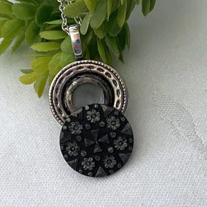 May include: A silver-toned pendant necklace with a circular design. The pendant features a detailed, dark gray, circular charm with floral and geometric patterns. The necklace is displayed on a white surface with green foliage in the background.