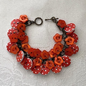 May include: A handmade bracelet featuring a mix of red buttons with white polka dots and flower-shaped buttons. The buttons are strung together with a dark metal chain and toggle clasp. A unique, whimsical accessory.