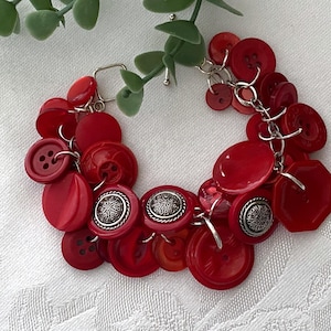 May include: A handmade bracelet featuring a collection of red buttons in various shapes and sizes. The buttons are connected with silver-colored chains and jump rings. Some buttons have a silver filigree design. The bracelet is displayed on a white surface.