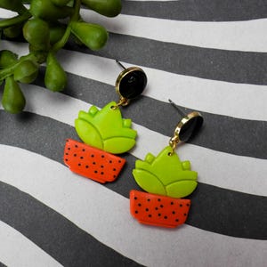 May include: A pair of dangle earrings featuring a lime green cactus design atop an orange pot with black polka dots. The earrings have black and gold accents and are set against a black and white striped background.