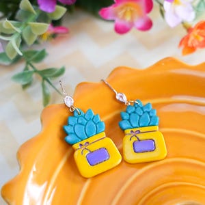 May include: A pair of handmade earrings featuring a succulent design. Each earring has a yellow pot with a purple rectangle and a turquoise succulent on top. The earrings are on a bright orange plate with flowers in the background.
