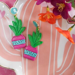 May include: A pair of handmade earrings featuring a miniature potted plant design. The earrings have green leaves and pink pots with blue accents. The earrings are displayed on a pink and white striped plate with gold trim.