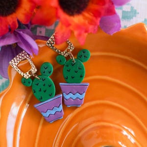 May include: A pair of cactus-shaped earrings with green cacti and purple pots with blue accents. The earrings are set against an orange dish with flowers in the background. The earrings have gold-colored square tops.