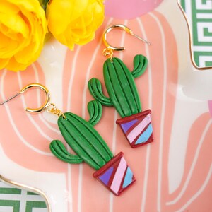 May include: A pair of gold hoop earrings with green cactus pendants. The cactus earrings are in a pot with red, purple, and blue stripes. The earrings are displayed on a pink and white striped plate with yellow roses.