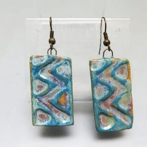 May include: A pair of handmade ceramic earrings. Each earring is a rectangular shape with a textured surface. The earrings feature a blue, white, orange, and purple color palette. The ear wires are a bronze color.