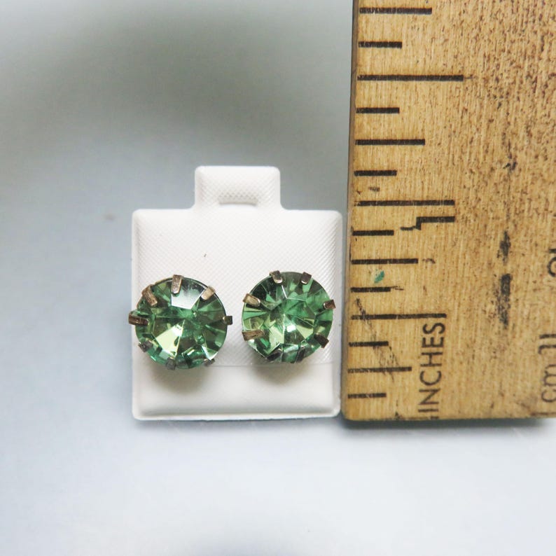 May include: A pair of green glass earrings with a silver setting. The earrings are displayed on a white card with a ruler for size reference.