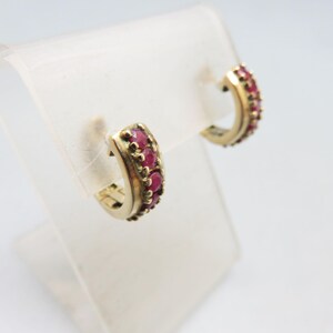 May include: A pair of gold-toned hoop earrings, each adorned with a row of vibrant red gemstones. The earrings are displayed on a clear, angled stand against a white background, showcasing their elegant design and color contrast.