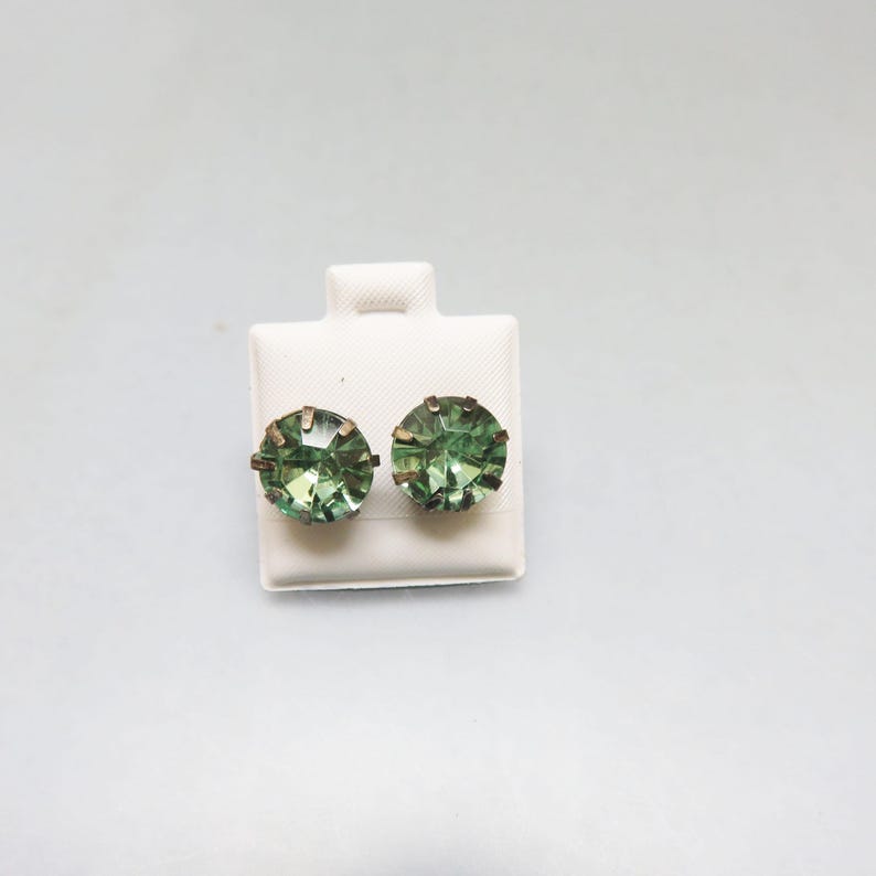 May include: A pair of green gemstone earrings with gold settings. The earrings are displayed on a white card.