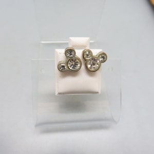 Pode incluir: Um par de brincos de press&atilde;o de prata com pedras de cristal transparentes em forma de cabe&ccedil;a do Mickey Mouse.