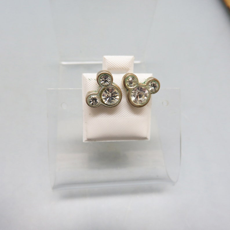 Pode incluir: Um par de brincos de press&atilde;o de prata com pedras de cristal transparentes em forma de cabe&ccedil;a do Mickey Mouse.