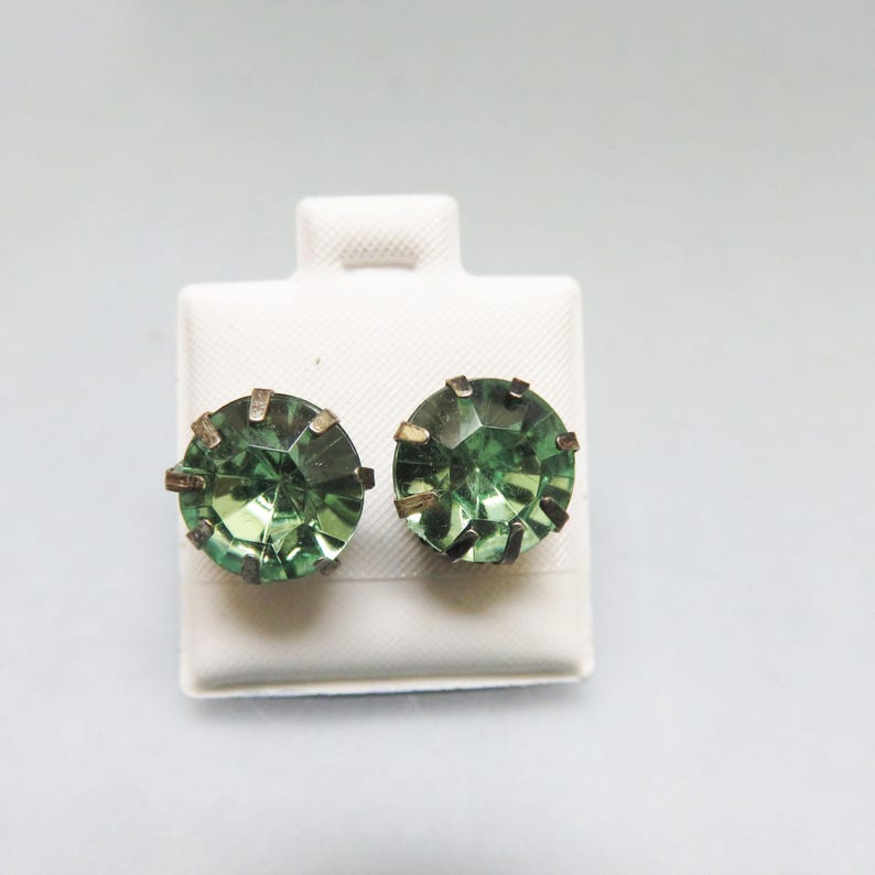 May include: A pair of green glass earrings with a gold-toned metal setting. The earrings are displayed on a white card.