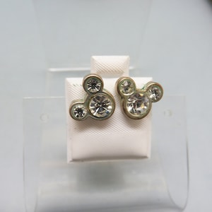 Pode incluir: Um par de brincos de press&atilde;o dourados com um design de silhueta do Mickey Mouse com strass transparentes.