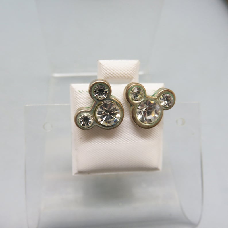 Pode incluir: Um par de brincos de press&atilde;o dourados com um design de silhueta do Mickey Mouse com strass transparentes.