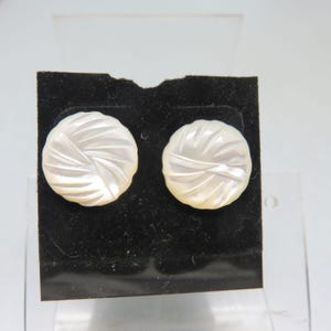May include: A pair of white, round earrings with a carved spiral design. The earrings are displayed on a black velvet surface. The earrings are made of a pearlescent material.