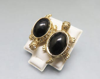 May include: A pair of gold-toned turtle-shaped earrings. Each turtle has a large, oval, black stone center, surrounded by small, clear crystals. The earrings are displayed on a white, textured surface.