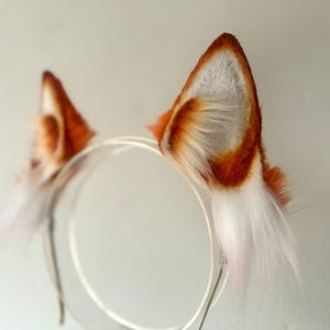 Op de afbeelding: Een paar oranje en witte nep bont kattenoren bevestigd aan een zilveren hoofdband. De oren zijn pluche en hebben een zachte, pluche textuur.