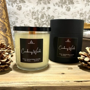 May include: Two black and white labeled jars of Cranberry Woods Coco-Apricot Wax Candle. The candle jars are on a wooden surface. The candle on the left is lit and has a clear glass jar. The candle on the right is in a black jar.