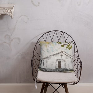 May include: A white throw pillow with a watercolor painting of a small white church with a cross on the roof. The church is surrounded by a light blue sky with green trees in the background. The pillow is sitting on a metal chair with a white cushion.