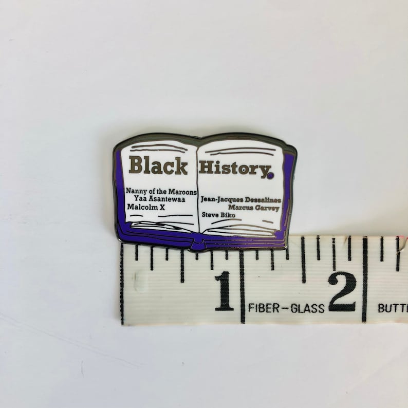 Op de afbeelding: Een zwarte en zilveren emaille pin in de vorm van een open boek met de woorden "Black History" op de omslag. Het boek vermeldt de namen van verschillende opmerkelijke zwarte figuren, waaronder Nanny of the Maroons, Yaa Asantewaa, Malcolm X, Jean-Jacques Dessalines, Marcus Garvey en Steve Biko.