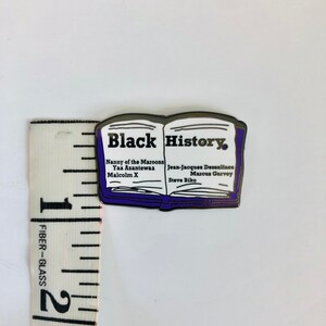 Op de afbeelding: Een zilveren emaille pin in de vorm van een open boek met de woorden "Black History" op de omslag. Het boek vermeldt de namen van verschillende prominente zwarte figuren, waaronder Nanny of the Maroons, Yaa Asantewaa, Malcolm X, Jean-Jacques Dessalines, Marcus Garvey en Steve Biko.