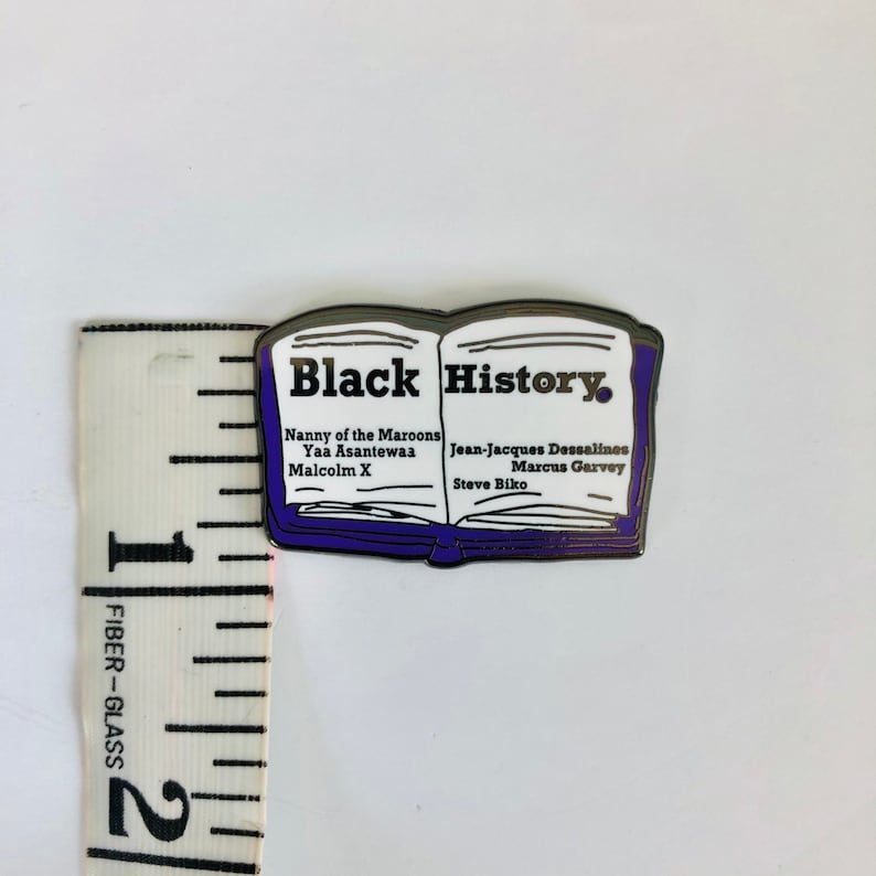 Op de afbeelding: Een zilveren emaille pin in de vorm van een open boek met de woorden "Black History" op de omslag. Het boek vermeldt de namen van verschillende prominente zwarte figuren, waaronder Nanny of the Maroons, Yaa Asantewaa, Malcolm X, Jean-Jacques Dessalines, Marcus Garvey en Steve Biko.