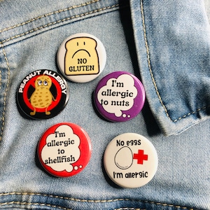 May include: Five round button pins with allergy information. The pins are on a blue denim background. The pins are white, black, purple, red, and white with a red cross. The text on the pins reads: "Peanut Allergy", "No Gluten", "I'm allergic to nuts", "I'm allergic to shellfish", and "No eggs I'm allergic".