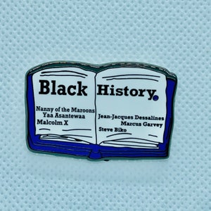 Op de afbeelding: Een blauwe en zilveren emaille pin in de vorm van een open boek met de woorden "Black History" op de omslag. Het boek vermeldt de namen van verschillende prominente zwarte figuren, waaronder Nanny of the Maroons, Yaa Asantewaa, Malcolm X, Jean-Jacques Dessalines, Marcus Garvey en Steve Biko.