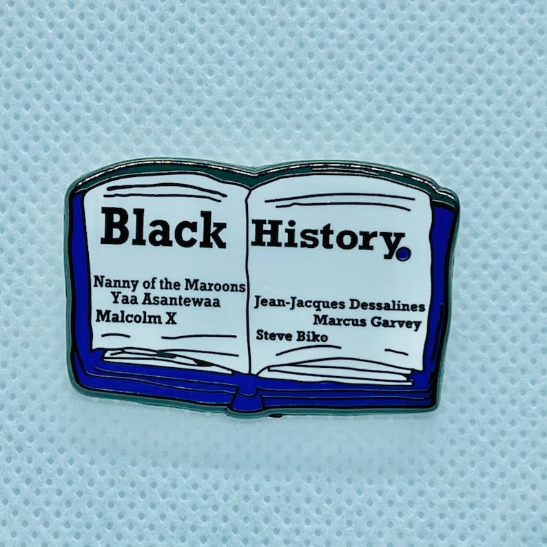 Op de afbeelding: Een blauwe en zilveren emaille pin in de vorm van een open boek met de woorden "Black History" op de omslag. Het boek vermeldt de namen van verschillende prominente zwarte figuren, waaronder Nanny of the Maroons, Yaa Asantewaa, Malcolm X, Jean-Jacques Dessalines, Marcus Garvey en Steve Biko.