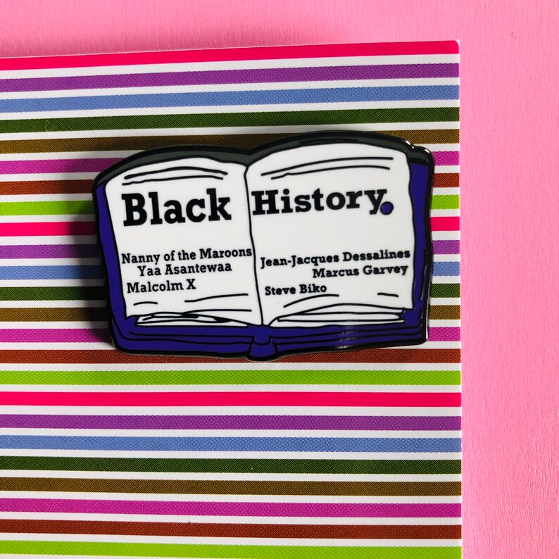 Op de afbeelding: Een blauwe en witte emaille pin in de vorm van een open boek met de tekst "Black History" op de omslag. Het boek vermeldt namen van belangrijke figuren in de zwarte geschiedenis, waaronder Nanny of the Maroons, Yaa Asantewaa, Malcolm X, Jean-Jacques Dessalines, Marcus Garvey en Steve Biko.