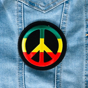 Puede incluir: Un parche negro con forma de símbolo de la paz con un borde rojo, amarillo y verde. El parche está cosido en una chaqueta vaquera azul.