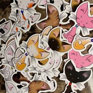 May include: A collection of cat stickers in various colors, including white, orange, brown, pink, and black. Each sticker features a cartoon cat with large ears, whiskers, and a small collar. The stickers are on a white background.