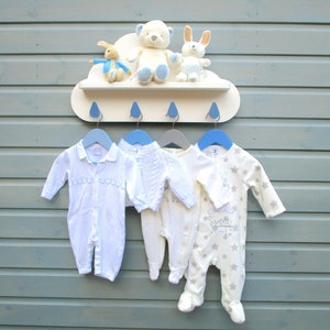 May include: A white cloud-shaped shelf with four blue hooks, holding four baby onesies. The onesies are white, cream, and light blue with different patterns. There are three stuffed animals on the shelf: a white bear, a white rabbit, and a brown rabbit.
