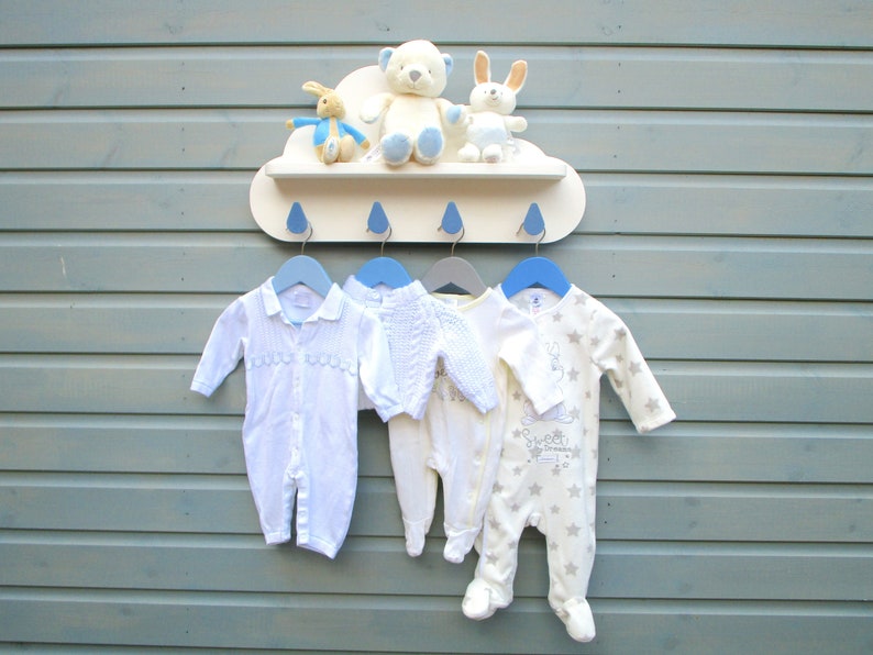 May include: A white cloud-shaped shelf with four blue hooks, holding four baby onesies. The onesies are white, cream, and light blue with different patterns. There are three stuffed animals on the shelf: a white bear, a white rabbit, and a brown rabbit.