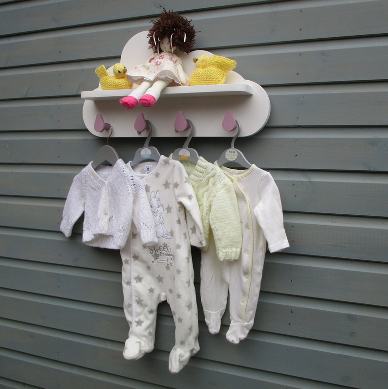May include: White cloud-shaped wall shelf with four hooks and two yellow bird figurines. The shelf has a doll with a pink dress and a white knitted cardigan hanging on the hooks.