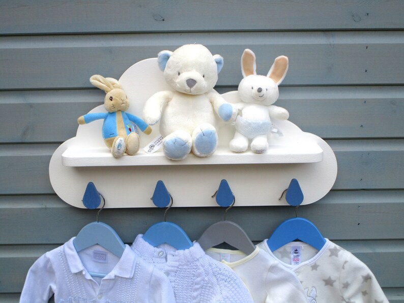 May include: White cloud-shaped shelf with blue raindrop hooks and a white teddy bear, a white bunny, and a Peter Rabbit plush toy on the shelf.  Four blue and gray hangers are hanging from the hooks.