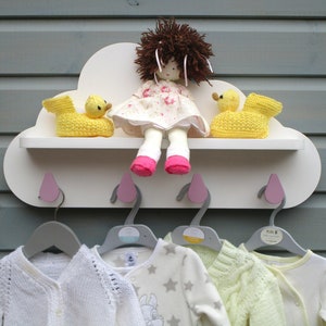 May include: A white cloud-shaped shelf with pink hooks and four gray hangers. The shelf has two yellow duck toys and a doll with brown hair and a pink dress. There are four baby clothes hanging on the hangers.