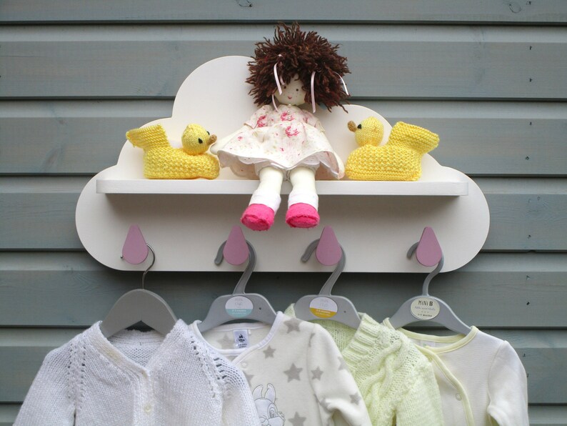 May include: A white cloud-shaped shelf with pink hooks and four gray hangers. The shelf has two yellow duck toys and a doll with brown hair and a pink dress. There are four baby clothes hanging on the hangers.