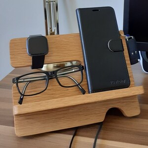 May include: A wooden phone stand with a black phone case, a black smartwatch, and a pair of eyeglasses. The stand has a cutout for the phone and a small shelf for the other items.