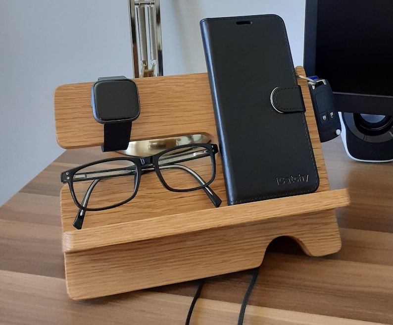 May include: A wooden phone stand with a black phone case, a black smartwatch, and a pair of eyeglasses. The stand has a cutout for the phone and a small shelf for the other items.