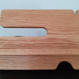 May include: A wooden phone stand with a cutout for a phone and a shelf for charging cables. The stand is made of light brown wood with a natural grain.