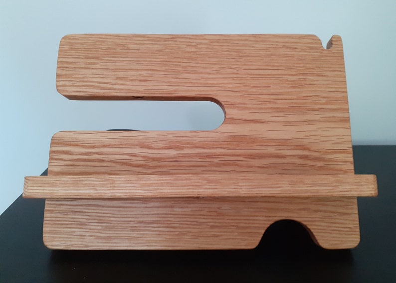 May include: A wooden phone stand with a cutout for a phone and a shelf for charging cables. The stand is made of light brown wood with a natural grain.