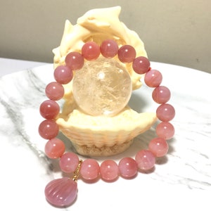May include: A pink agate gemstone bracelet with a shell charm. The bracelet is made of round beads and is strung on a red cord. The charm is made of a pink agate gemstone and is shaped like a shell.