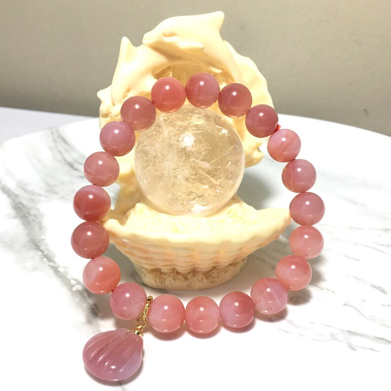 May include: A pink agate gemstone bracelet with a shell charm. The bracelet is made of round beads and is strung on a red cord. The charm is made of a pink agate gemstone and is shaped like a shell.